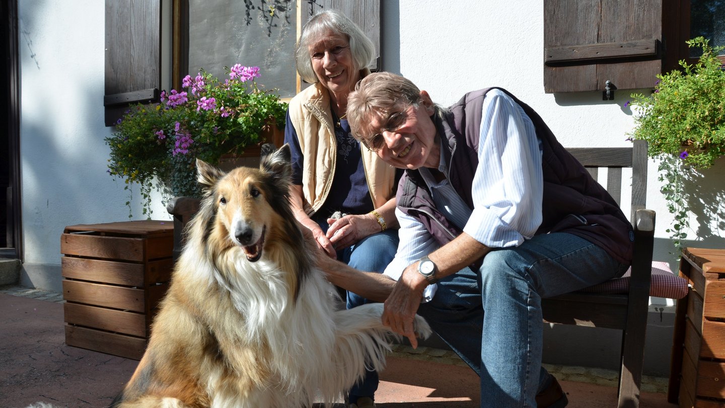 zwei ältere Menschen und ein Collie auf einer Bank vor dem Sonnenhof