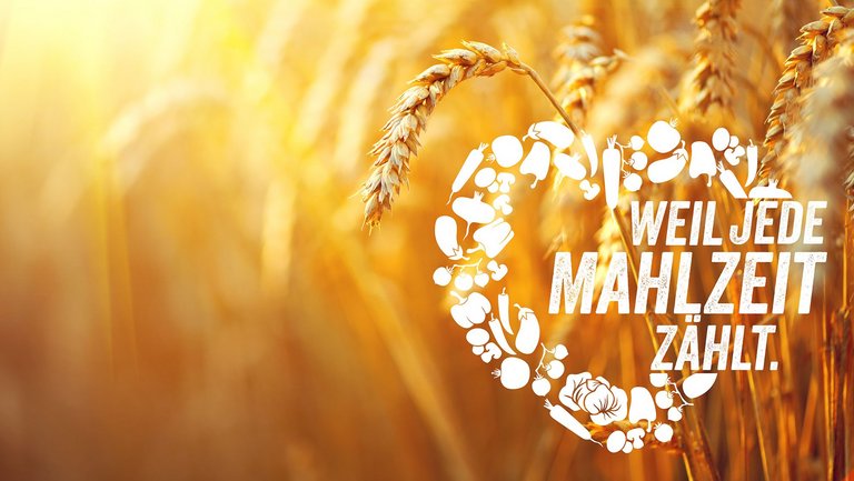 Pflanzlichen Lebensmittel formaen ein weißes Herz mit der Aufschrift "Weil jede Mahlzeit zählt" vor einer Gertreide-Ähre im Hintergrund