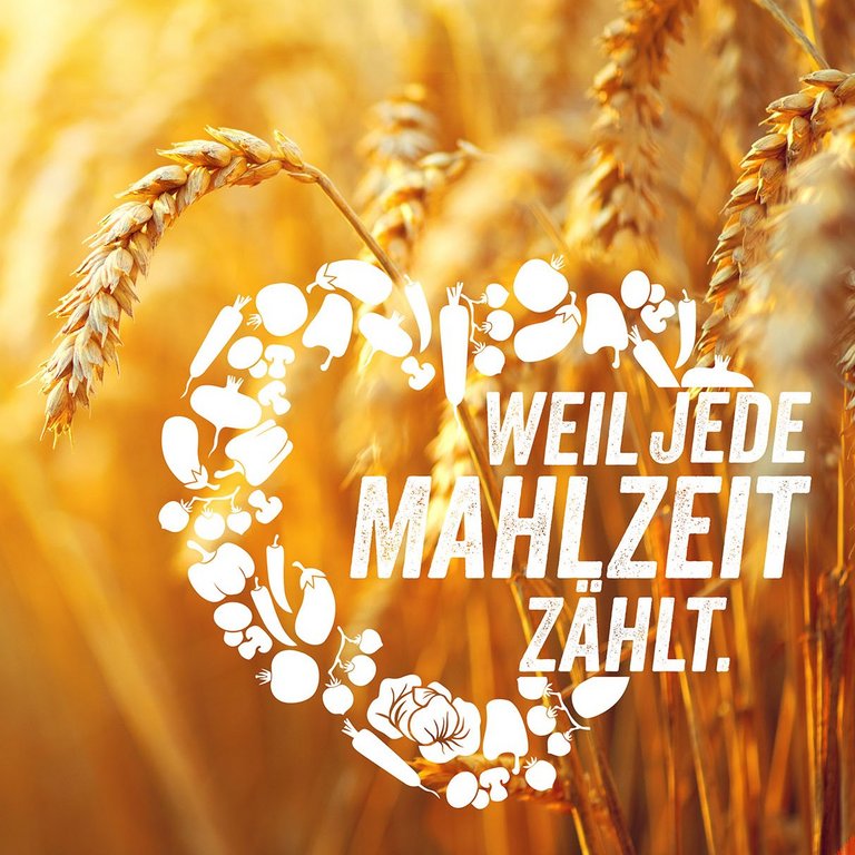 Pflanzlichen Lebensmittel formaen ein weißes Herz mit der Aufschrift "Weil jede Mahlzeit zählt" vor einer Gertreide-Ähre im Hintergrund